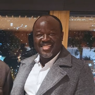 Smiling man wearing a gray coat and white shirt standing indoors with lights in the background.