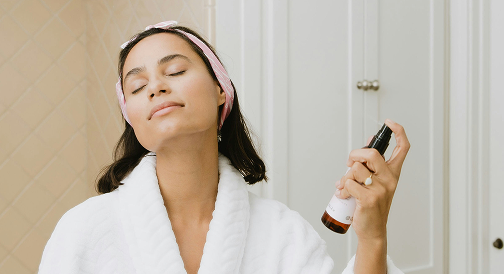 A woman in her robe with her hair pulled back spritzing toner on her face
