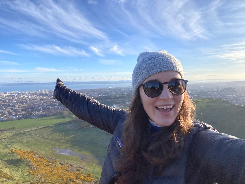 Traveler smiling and pointing toward panoramic views of Edinburgh, Scotland, from Arthur’s Seat on a clear blue-sky day