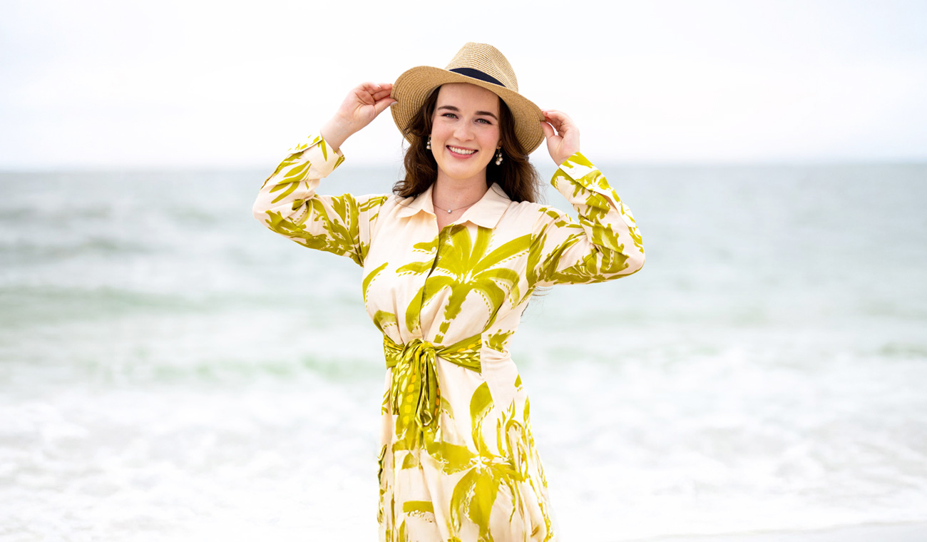 Woman smiling in a tropical palm print dress and straw hat on the beach with the ocean waves behind her, representing personal and adventurous travel with Moments by Meagan.
