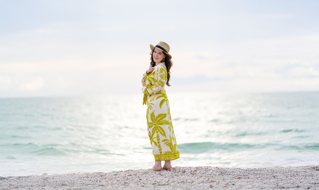 Woman in a tropical palm print dress and straw hat smiling barefoot on a seashell beach at sunset with the ocean glowing in the background, representing adventure travel with Moments by Meagan