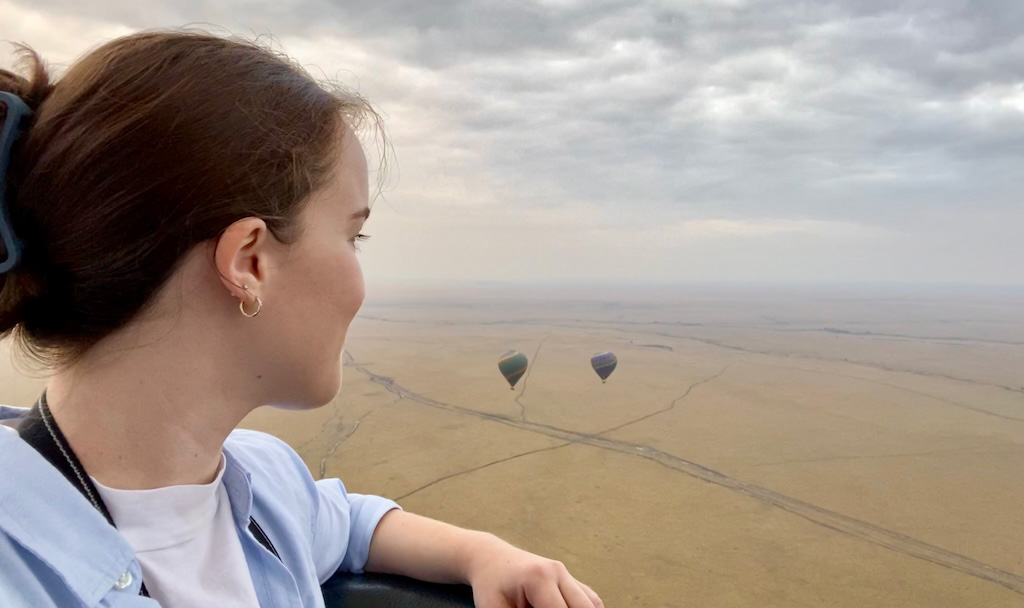 Traveler looking out from a hot air balloon ride over the African savanna with two colorful balloons floating in the distance under a dramatic cloudy sky, symbolizing adventure travel with Moments by Meagan