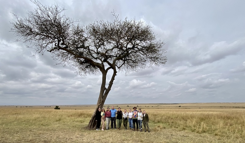 Travel group standing together beneath a lone acacia tree in the African savanna under a cloudy sky, highlighting the community and adventure of safari travel with Moments by Meagan