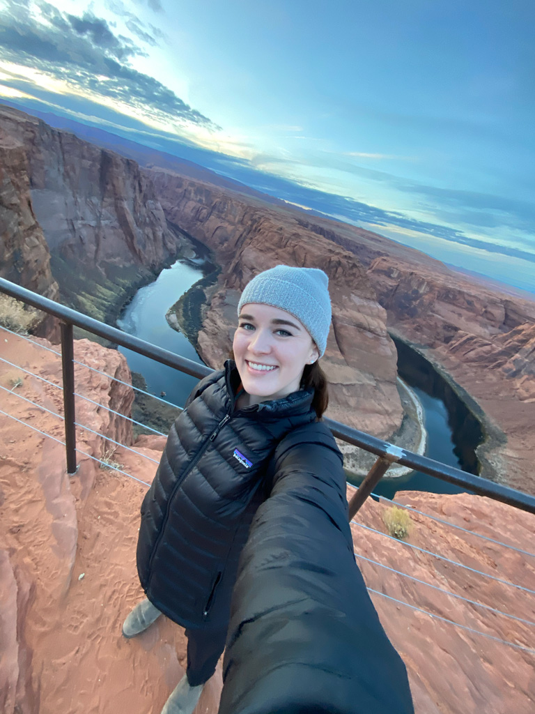 Traveler smiling in a winter jacket and beanie while taking a selfie at Horseshoe Bend in Arizona, with the Colorado River winding through dramatic red rock canyons at sunset.