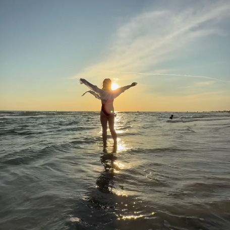 Traveler standing in the ocean at sunset with arms outstretched, silhouetted against the glowing sky and waves, capturing the freedom and joy of adventure travel with Moments by Meagan.