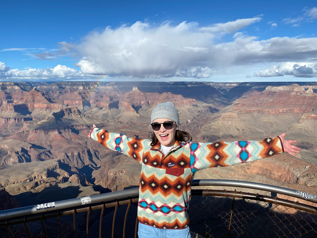 Traveler smiling with arms outstretched at a Grand Canyon overlook, wearing sunglasses and a colorful patterned sweater on a clear day with dramatic red rock cliffs and a rainbow in the distance.