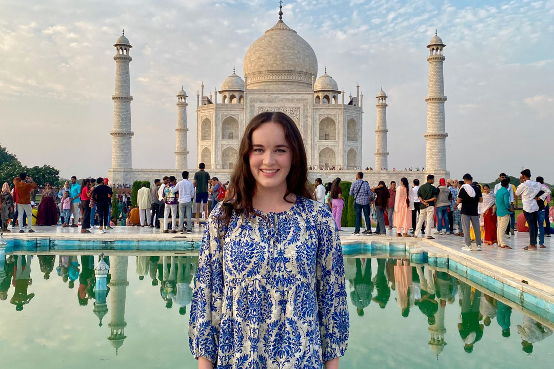 Traveler smiling in front of the Taj Mahal in Agra, India, wearing a blue patterned dress with the white marble monument and reflecting pool behind her, capturing the beauty of cultural and adventure travel.