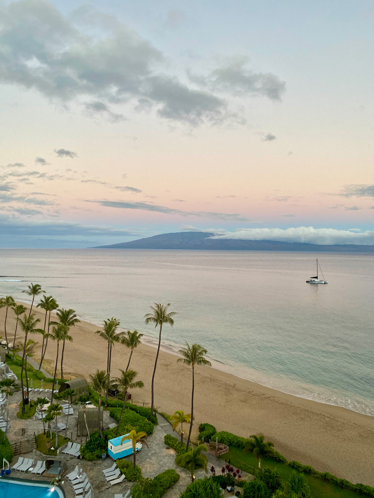 Peaceful sunset view of a sandy beach in Maui, Hawaii, with palm trees, calm ocean waves, and a sailboat anchored near the horizon.
