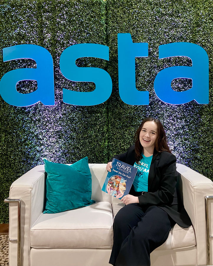 Smiling travel advisor sitting at the ASTA conference, holding a travel magazine while seated on a white couch with a green wall backdrop and large ASTA logo.
