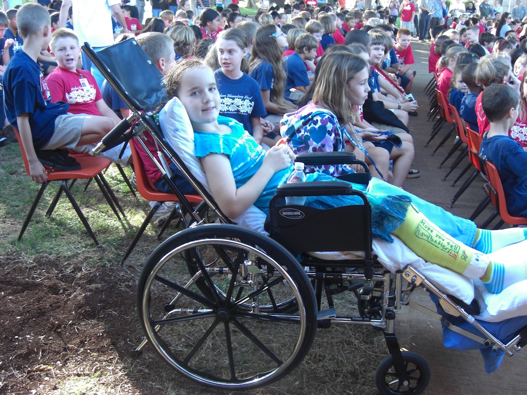 Young student in a wheelchair with a cast on her leg, sitting among classmates during a school assembly outdoors, highlighting accessibility and inclusion.