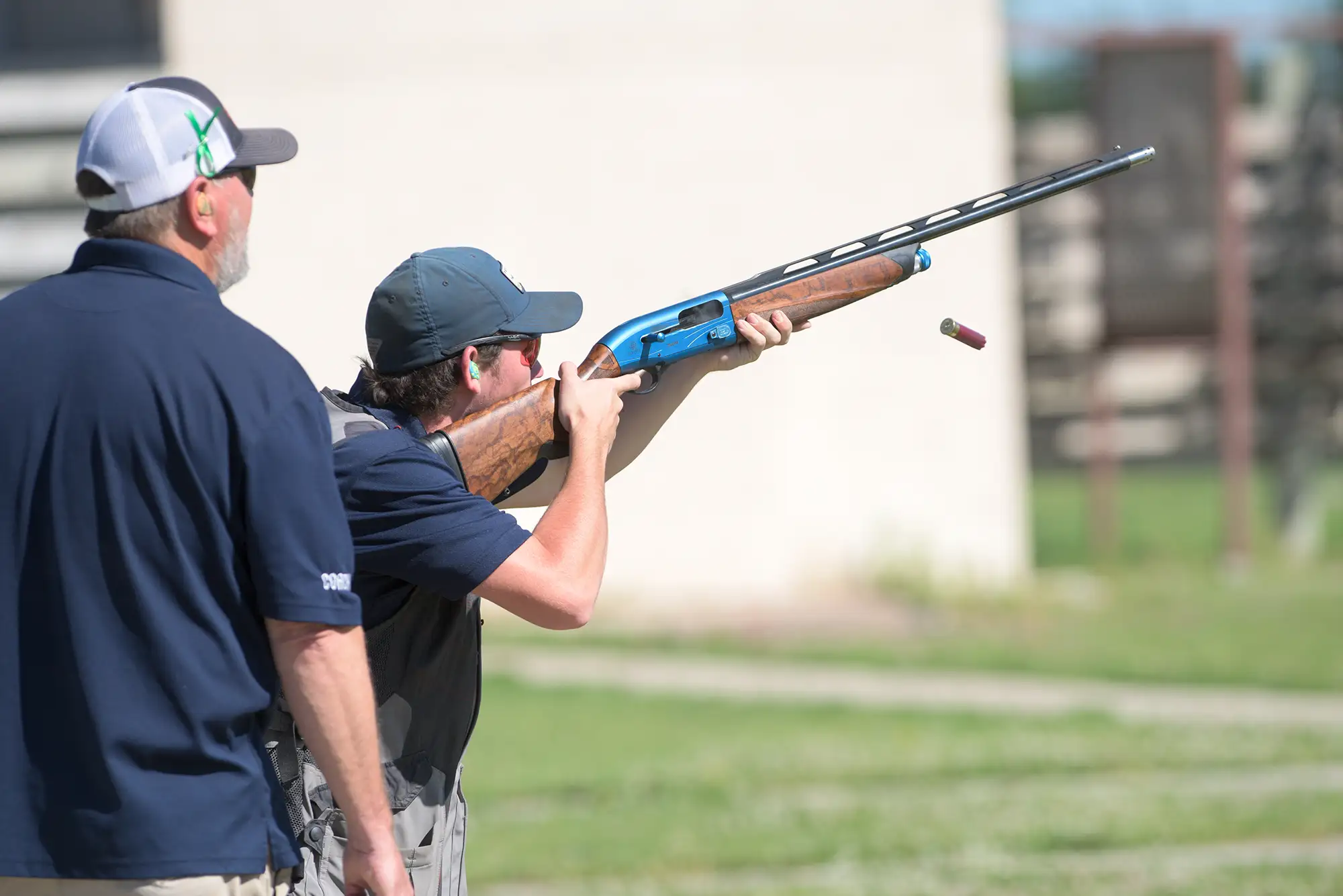 A person stands on a grassy field aiming a rifle, wearing a blue shirt and jeans. The sunny day and open sky convey a calm, focused atmosphere.