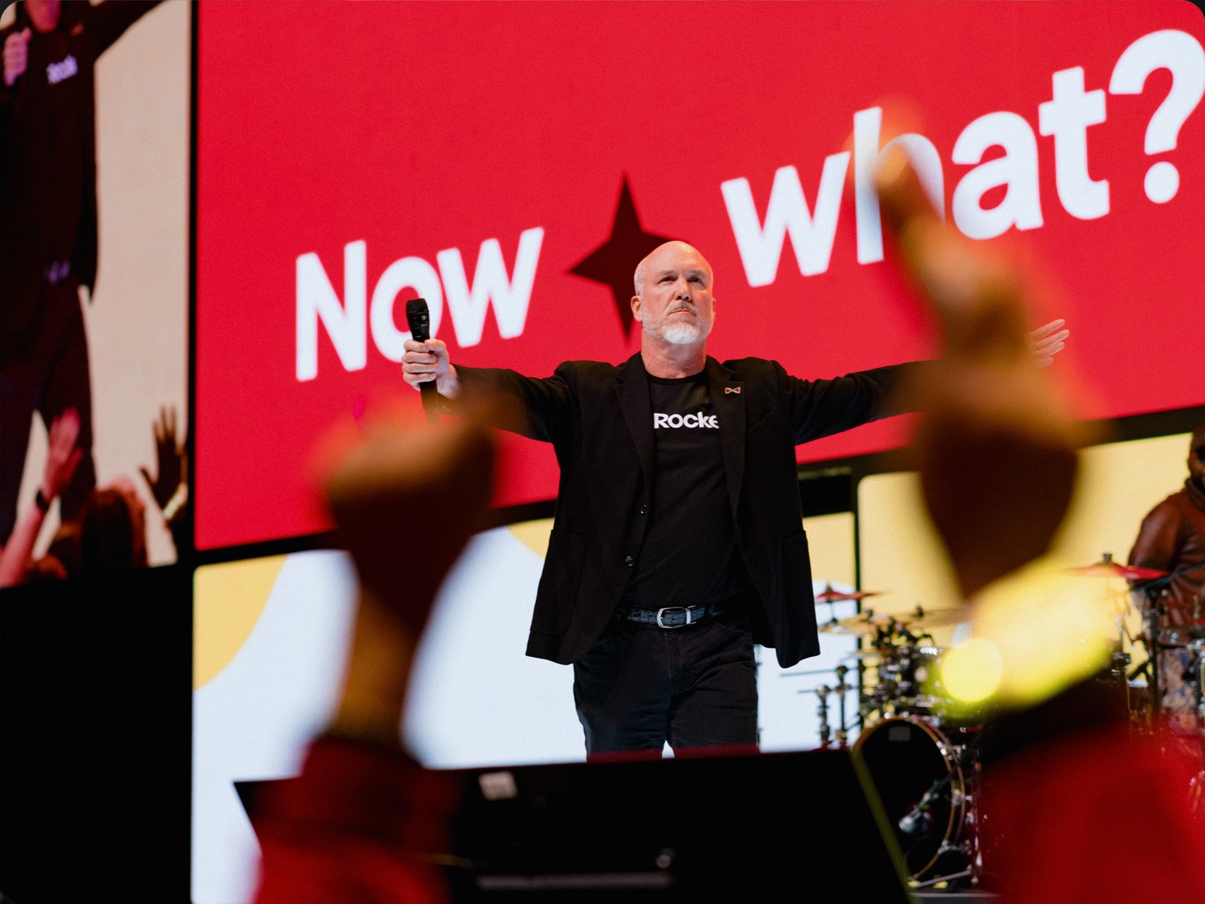 Bill Emerson stands on stage with arms outstretched holding a microphone, in front of a bright red screen displaying the words “Now what?” while the crowd cheers.