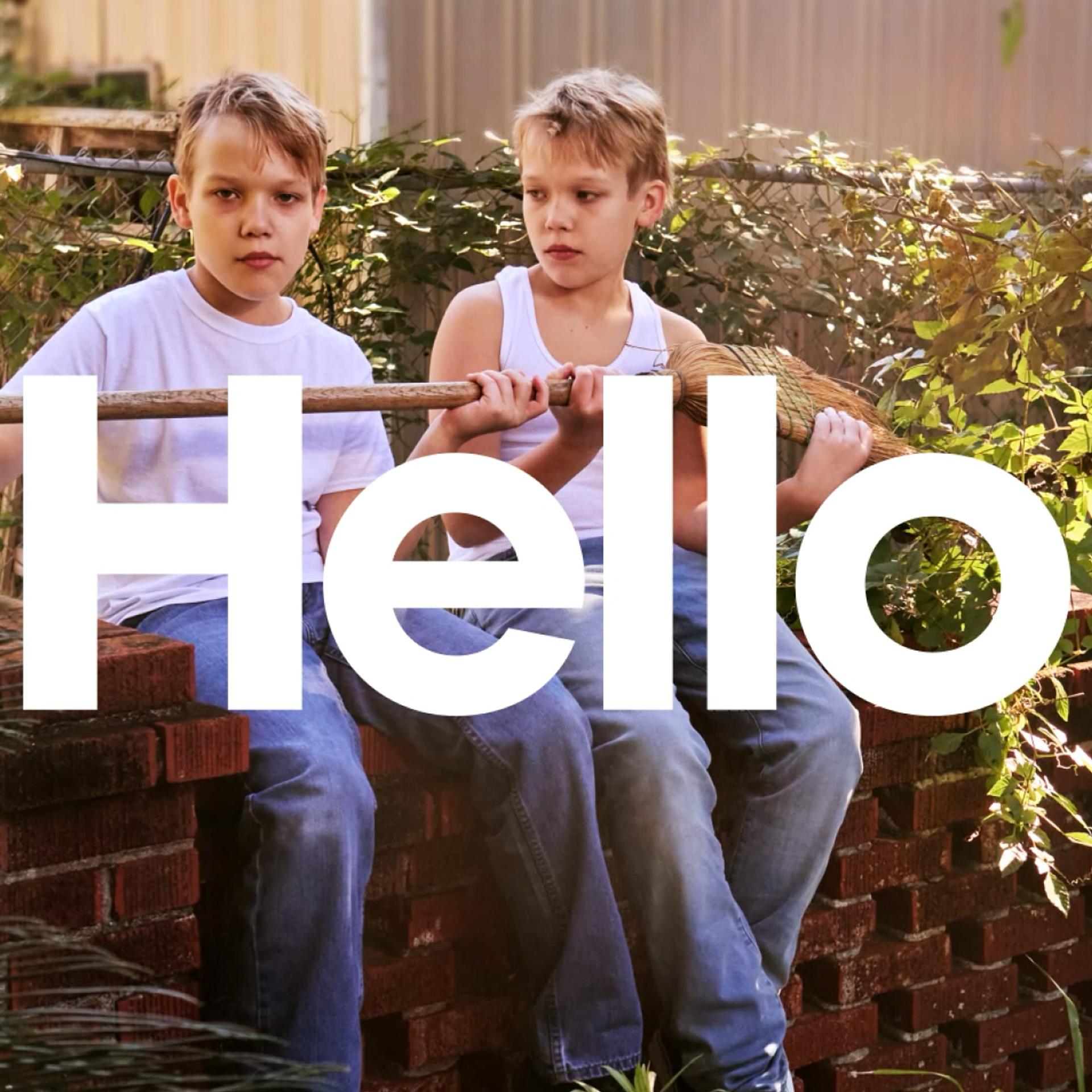 Two young boys in casual clothes sit on a low brick wall outside, holding a broom handle together. Large white text across the image reads "Hello."