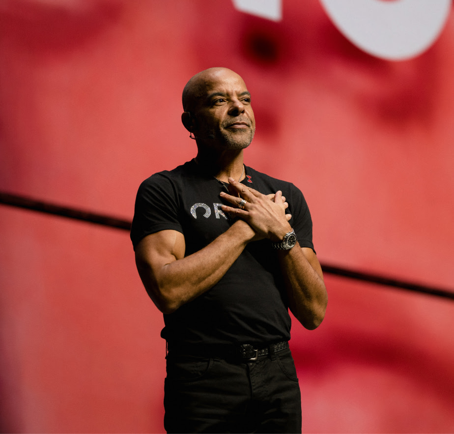 Jonathan Mildenhall stands on stage with his hands over his chest, wearing a black shirt with a Rocket logo, in front of a red background.