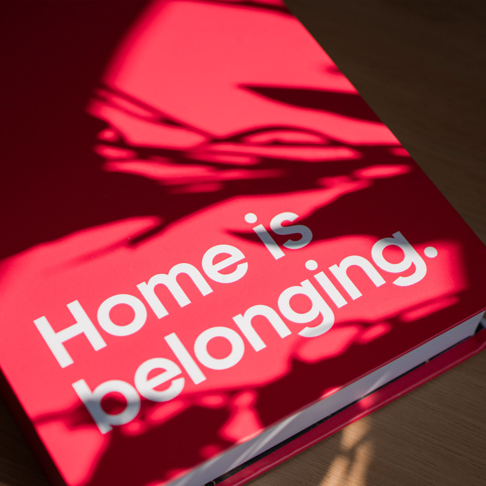 A red book cover on a wooden table with a shadow across it. White text reads: "Home is belonging."