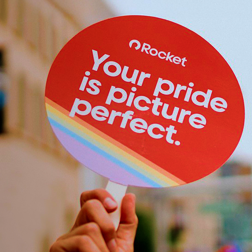 A person holds a round, red sign with a rainbow stripe. White text reads: "Your pride is picture perfect."