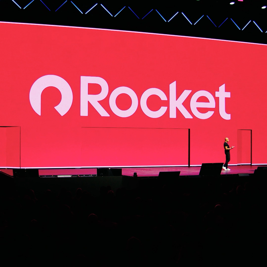A large presentation screen displays the Rocket logo in white on a bright red background. A speaker stands on stage in front of the screen addressing the audience.