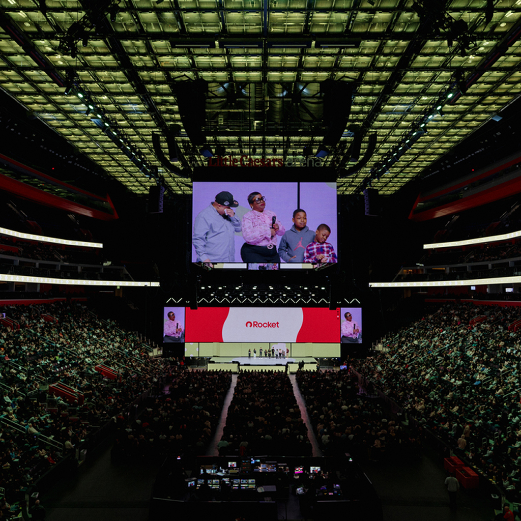 A large audience fills an arena as people on stage are shown on a massive screen. The screen displays the Rocket logo as a family shares their story during a company event.