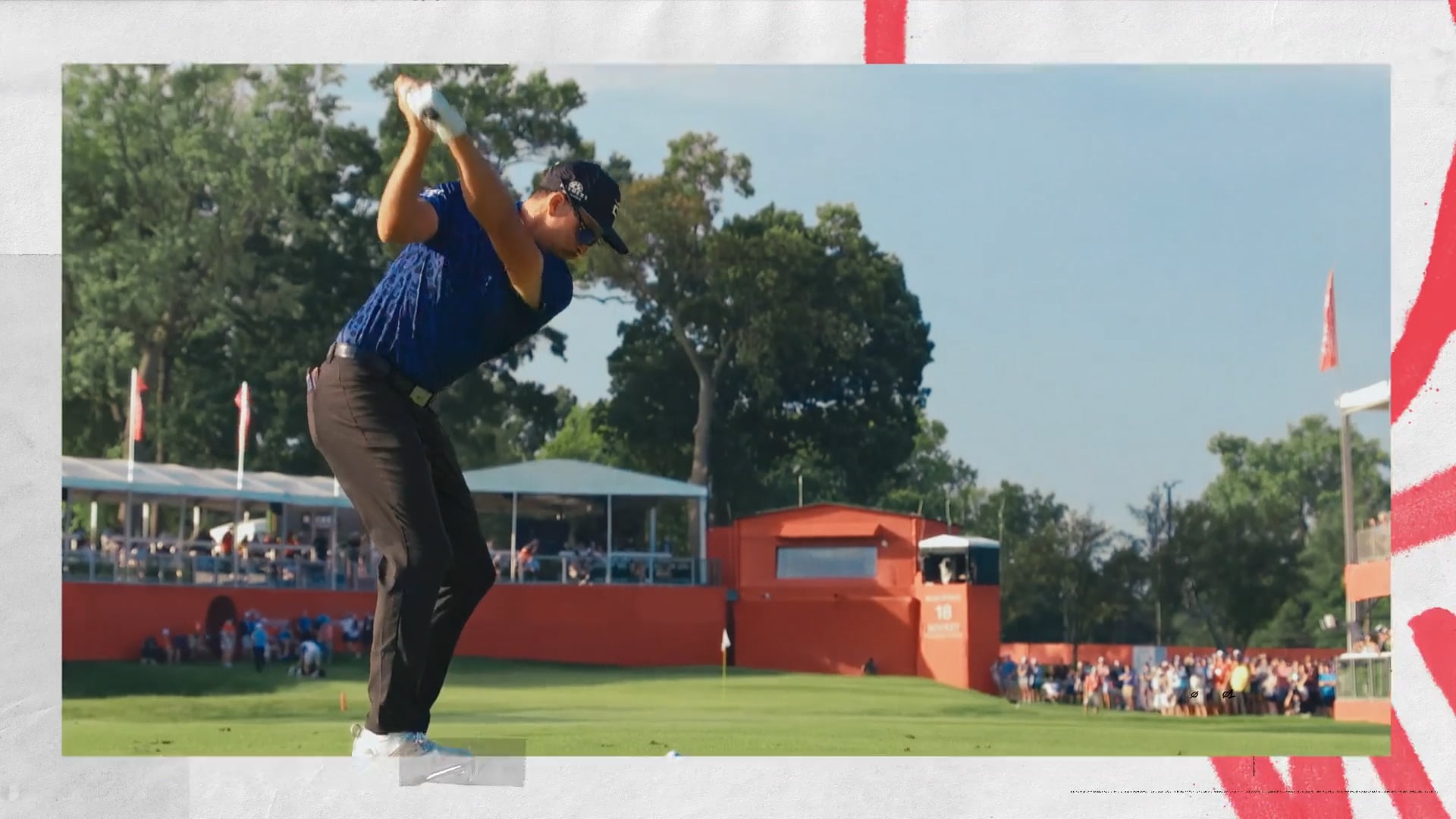 A golfer in a blue shirt and black pants takes a swing during a tournament, with spectators and red tournament structures in the background.