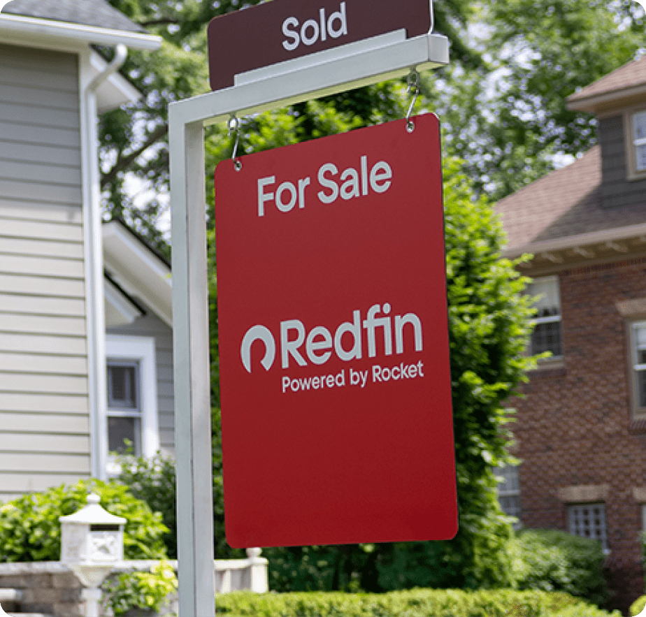 A red “For Sale” sign with the Redfin logo and “Powered by Rocket” text, hanging in front of a home with a “Sold” tag above it.