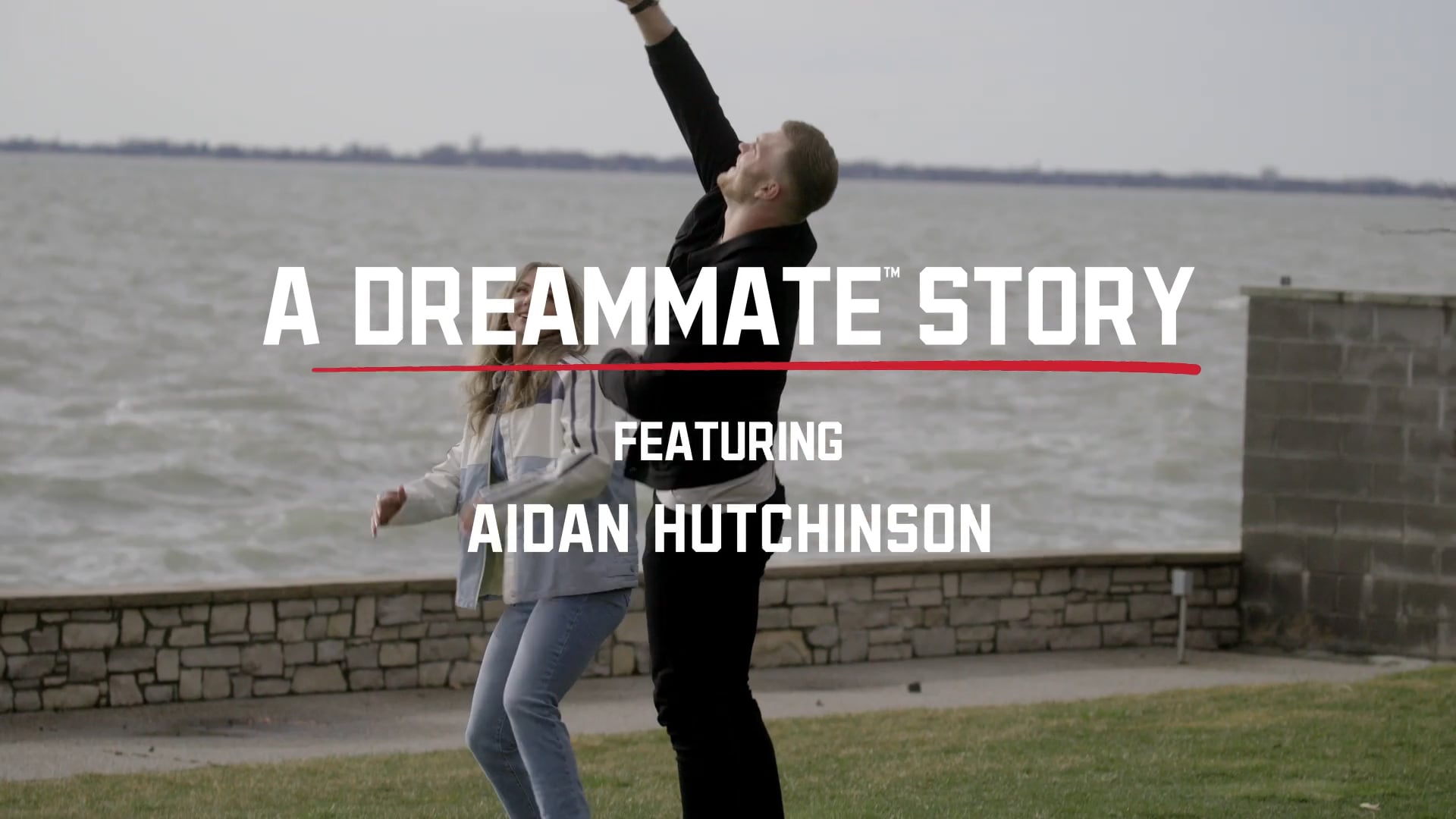 A man raises his arm triumphantly while standing with a woman by a waterfront, overlaid with text that reads: “A Dreammate Story featuring Aidan Hutchinson.”