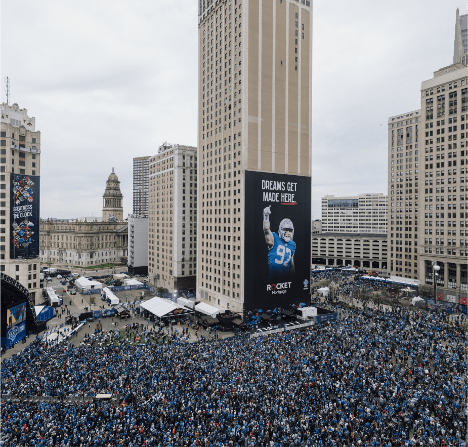 A massive crowd gathers in downtown Detroit for the NFL Draft, with tall buildings featuring banners, including one with a football player and the slogan “Dreams Get Made Here.”