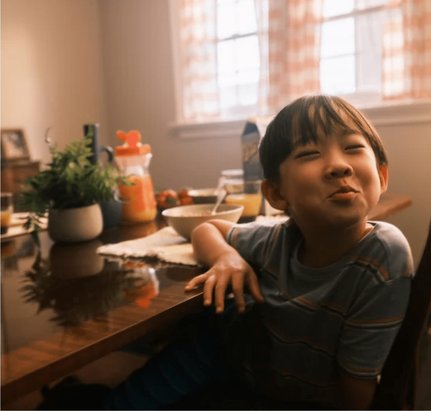 A young boy sits at a dining table with breakfast items, smiling mischievously while sunlight streams through curtained windows behind him.