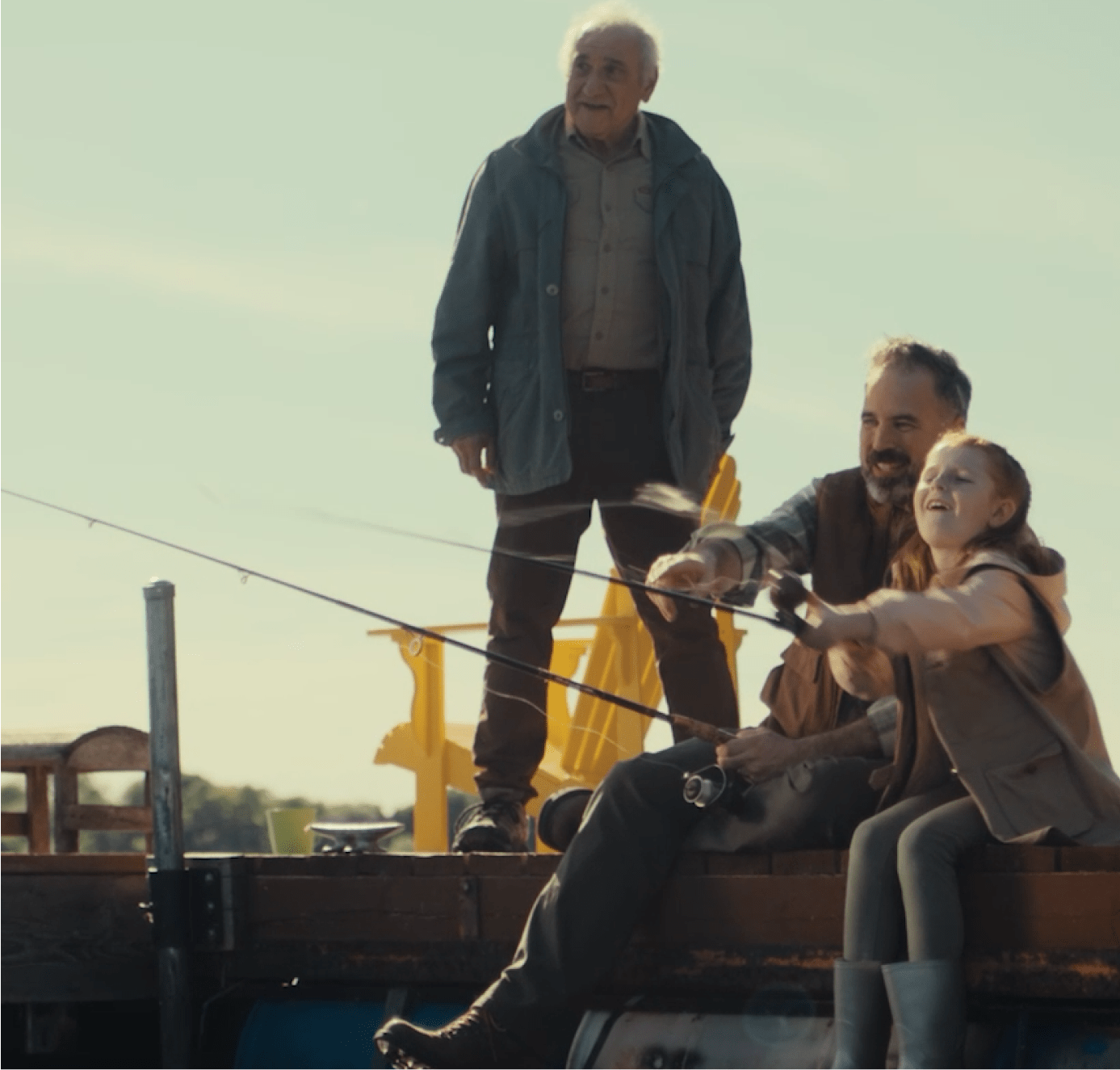 Three generations—an elderly man standing, a middle-aged man sitting, and a young girl—fish together on a dock under a clear sky.