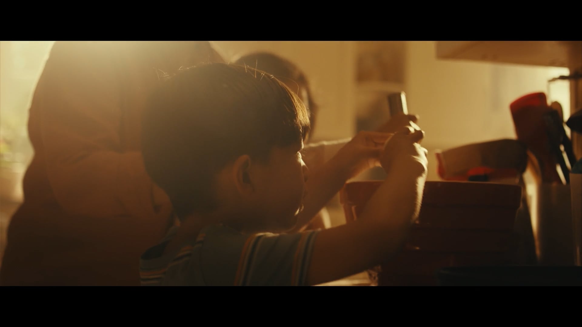 A boy helps prepare food in a warmly lit kitchen, holding a utensil while an adult stands beside him in the golden sunlight.