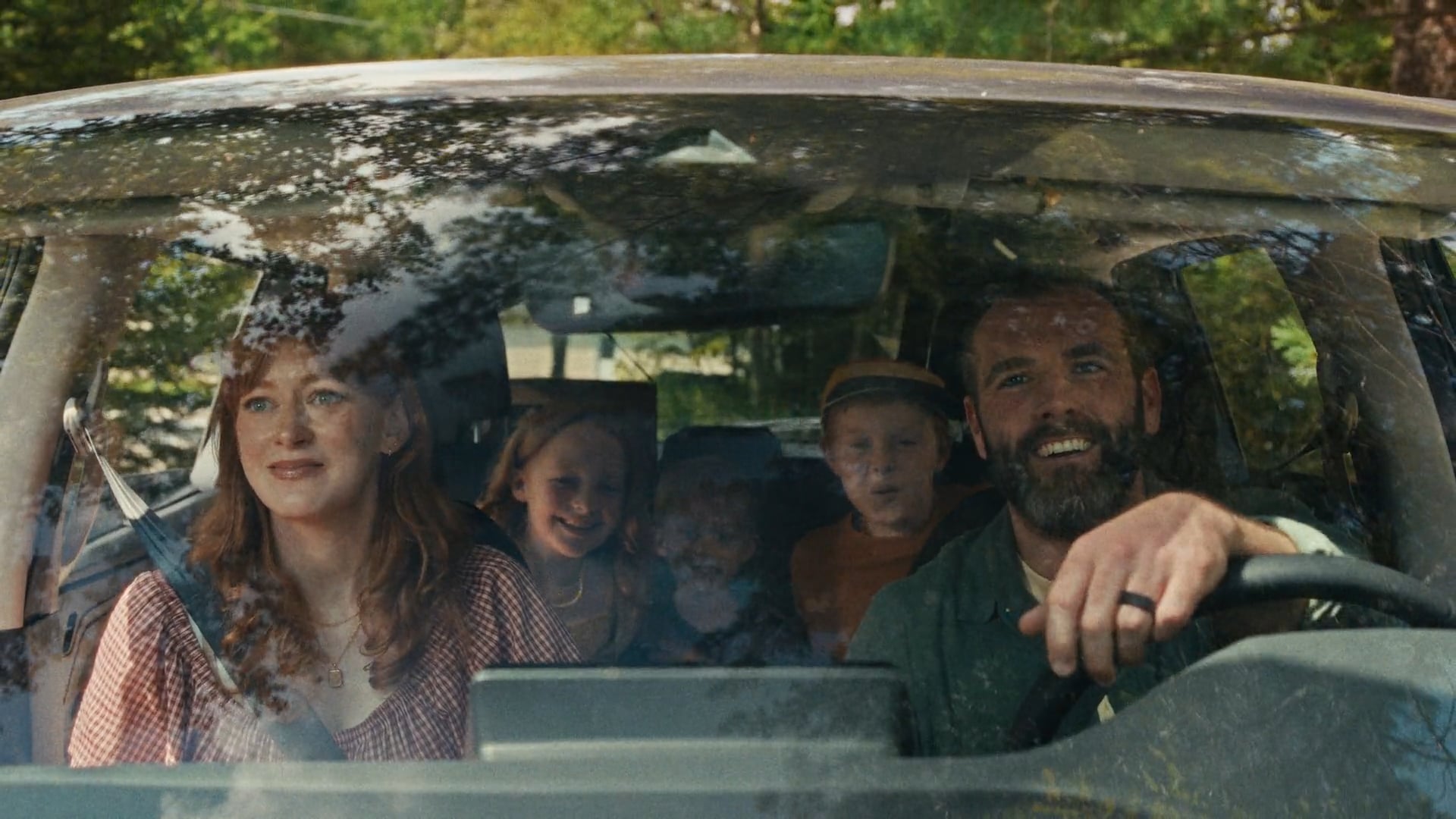 A family of five sits inside a car, seen through the windshield; two parents in front and three children smiling in the back seat.