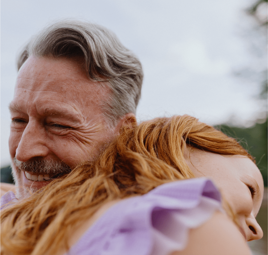 An older man with gray hair smiles warmly while hugging a young girl with red hair outdoors near a lake.