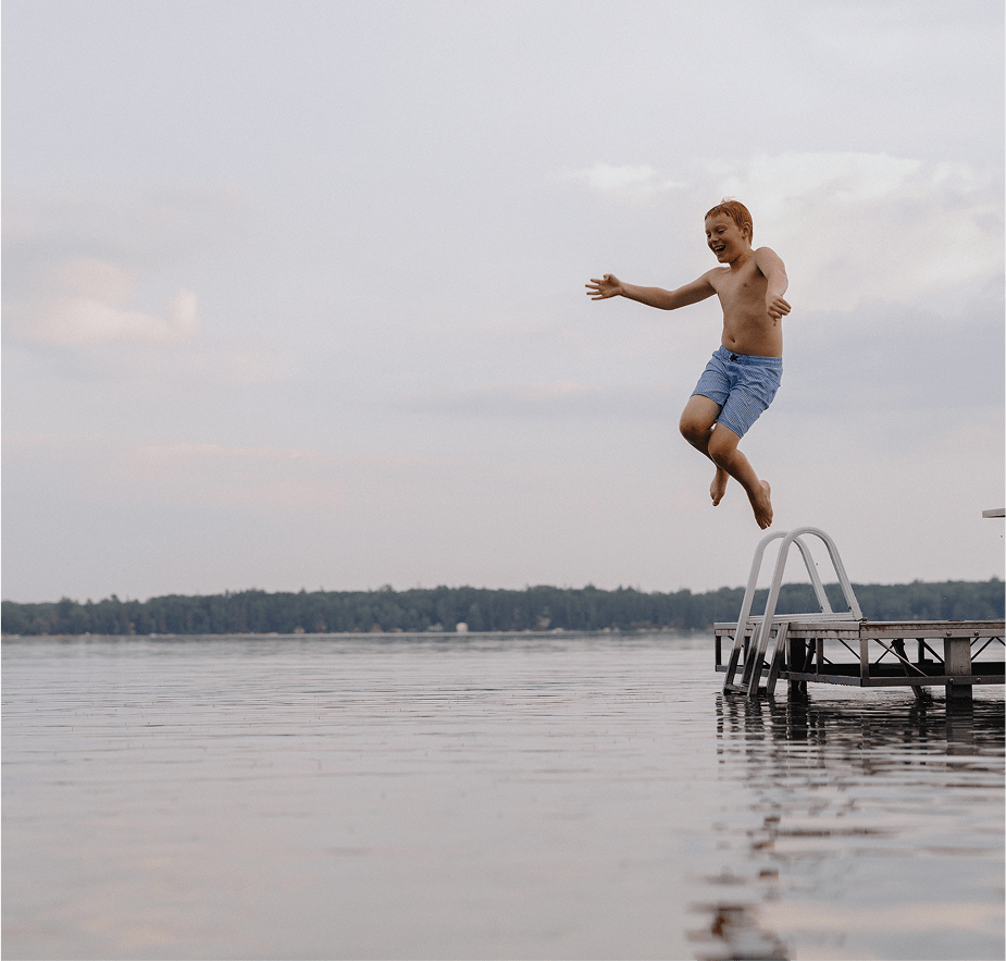 A boy in blue swim shorts jumps joyfully off a dock into a calm lake on a cloudy day.