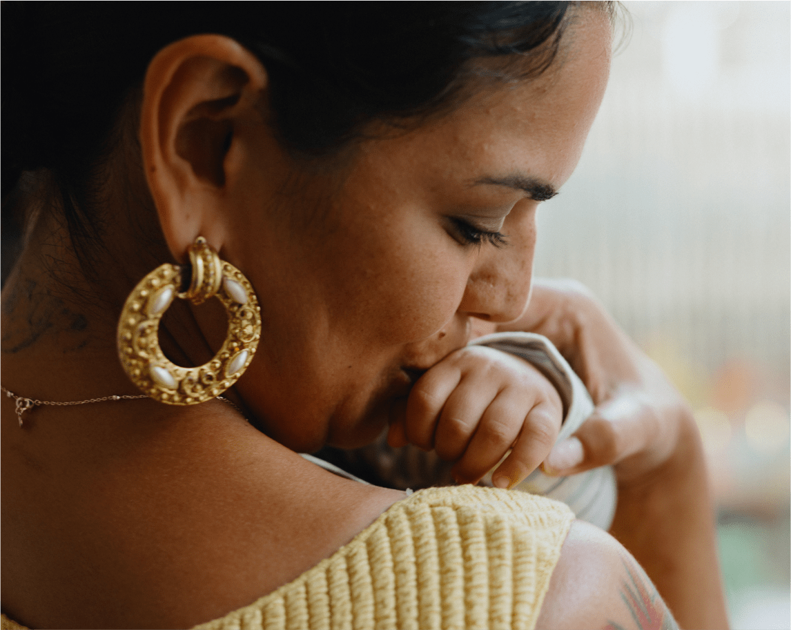 A woman with gold hoop earrings gently kisses the hand of a baby she is holding close.