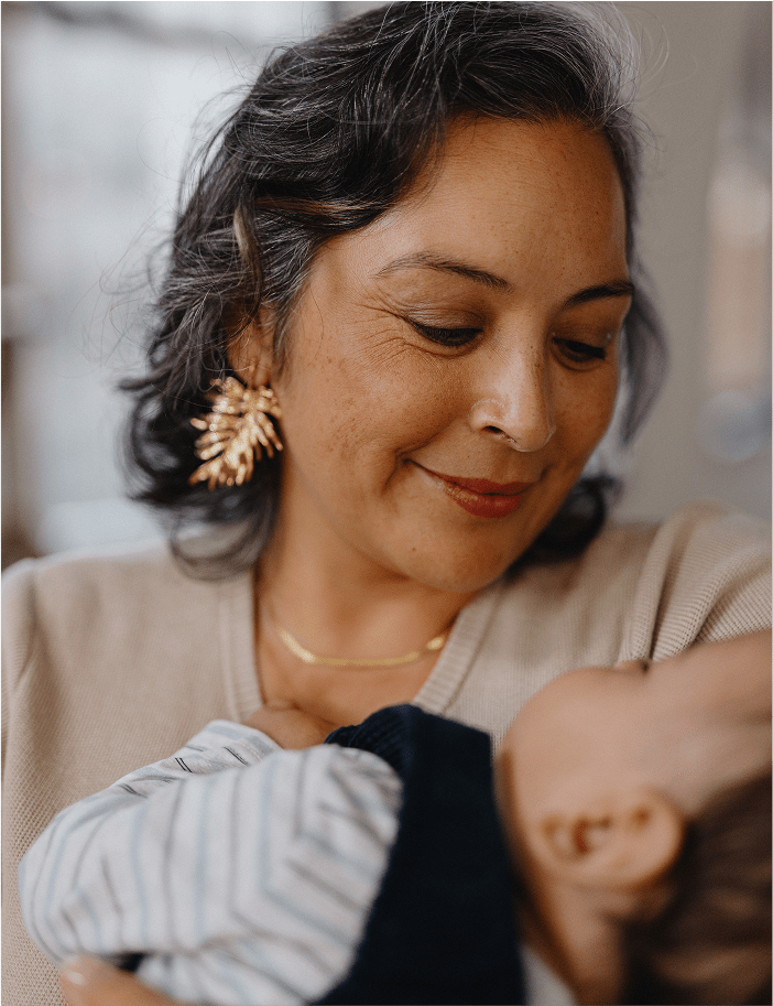 A woman with short gray-streaked hair smiles down at a baby in her arms, wearing a soft beige sweater and gold jewelry.
