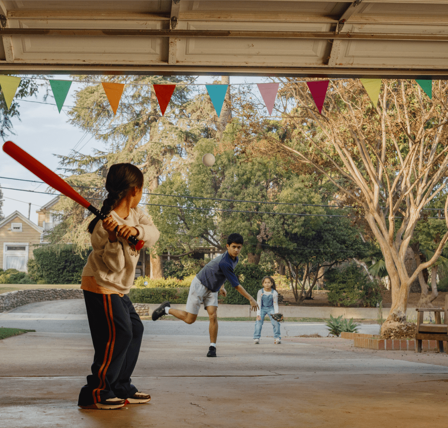 Three children playing baseball in a driveway, framed by a garage and colorful pennants. A girl in the foreground swings a red bat.