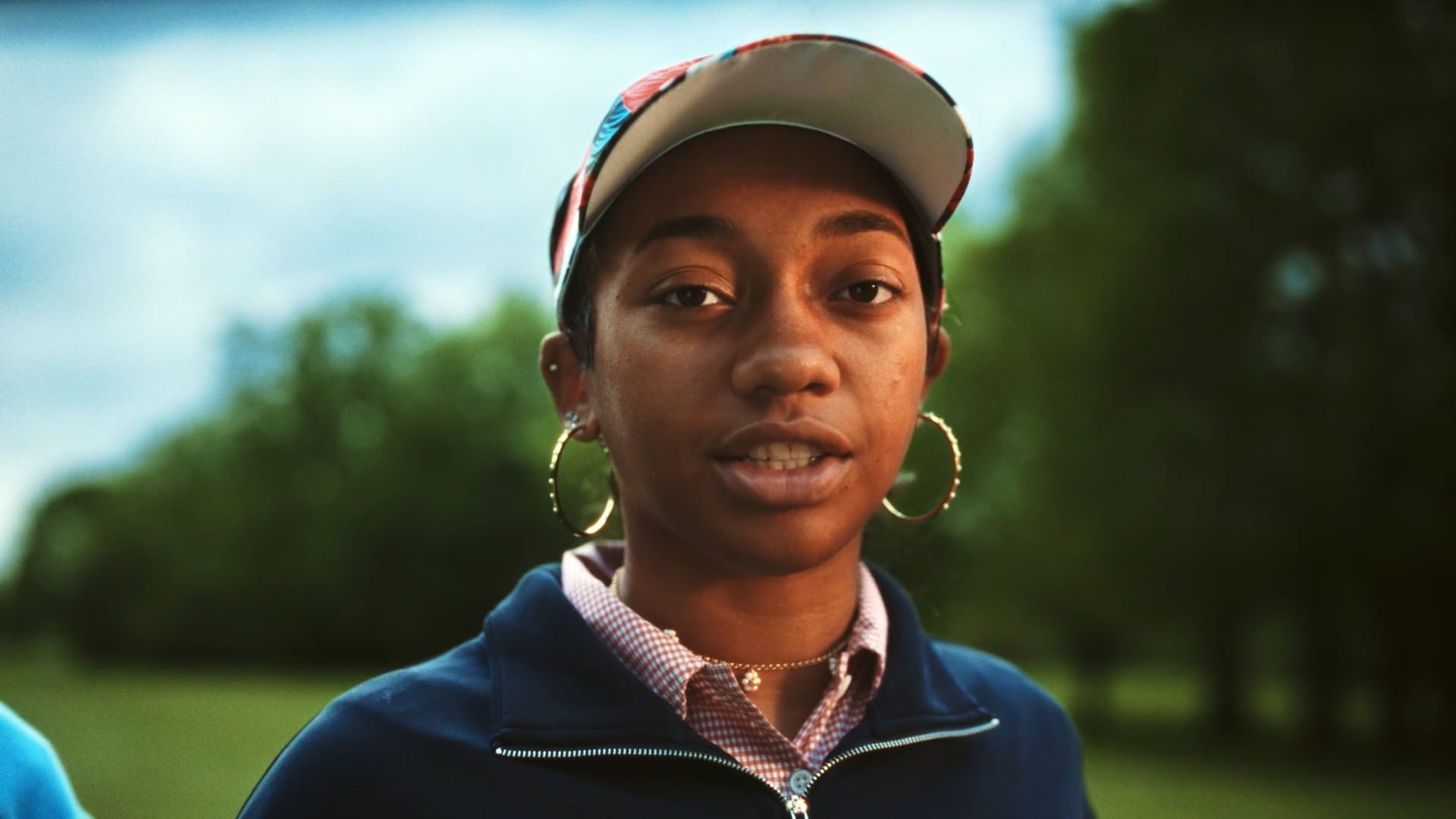 A young woman wearing a colorful cap, hoop earrings, and a blue jacket speaks outdoors on a golf course.
