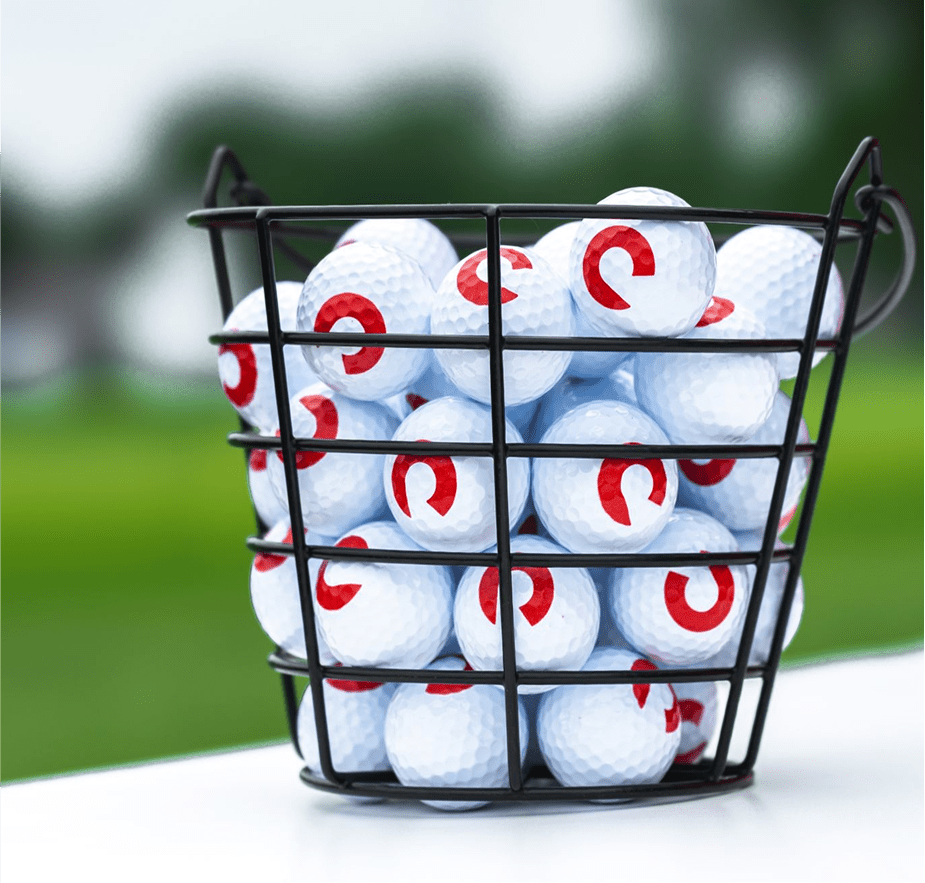 A black wire basket filled with white golf balls marked with a red circular logo, set on a golf course.