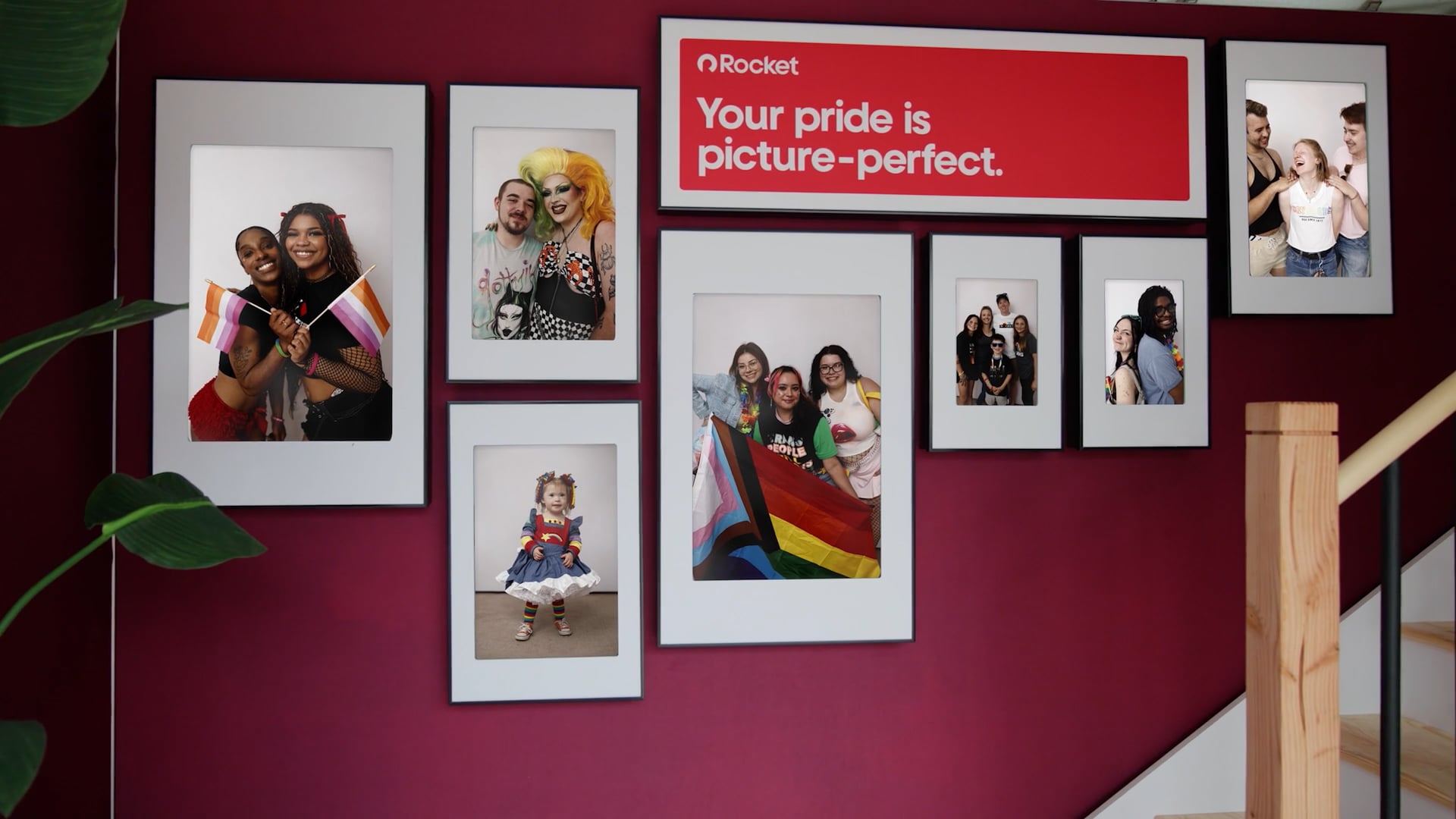 A gallery wall of framed photos on a red wall with a sign that reads, "Your pride is picture-perfect." The photos show various LGBTQ+ individuals and groups.