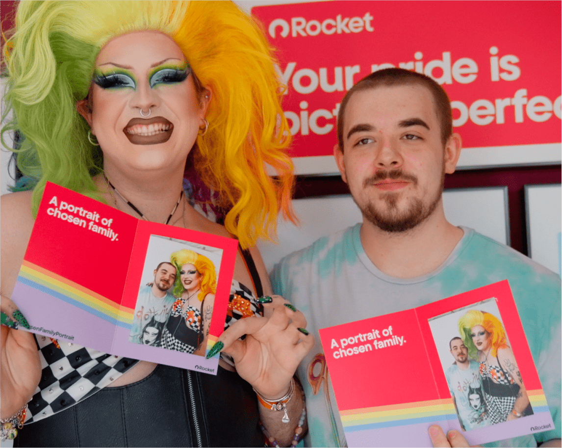 A drag queen with green and yellow hair and a young man smile while holding identical red cards that read "A portrait of chosen family."