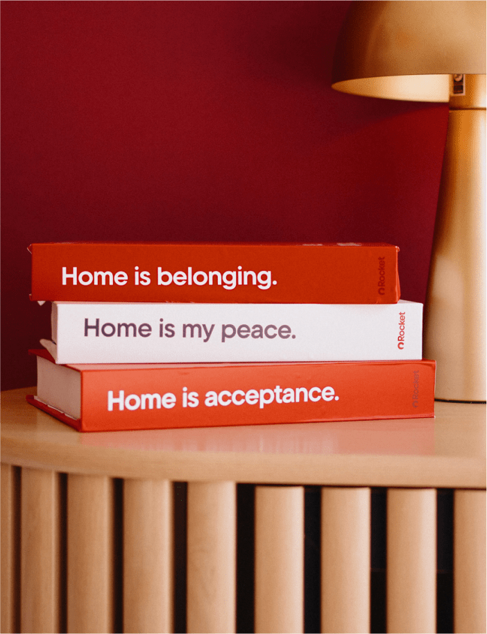 A stack of three books: two red and one white. The titles read: "Home is belonging," "Home is my peace," and "Home is acceptance."