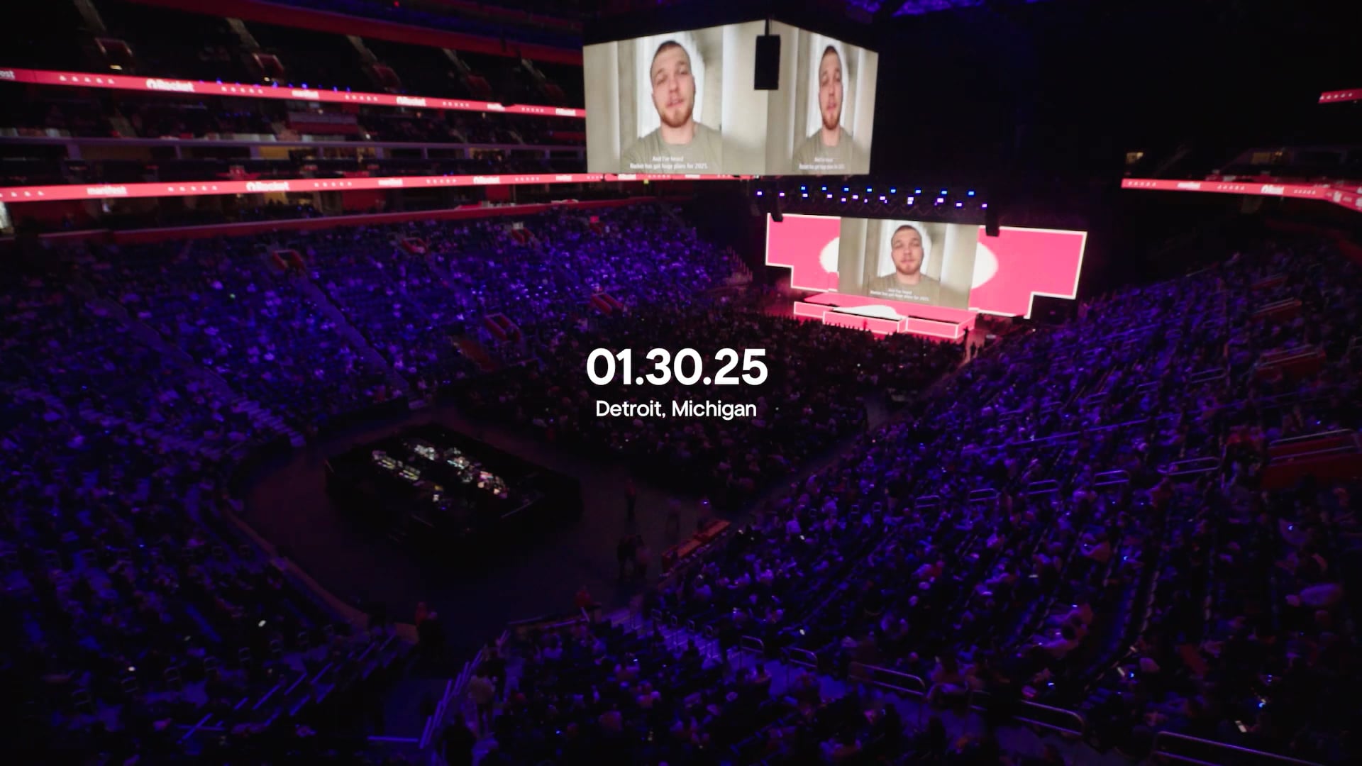 An arena filled with people illuminated by blue lighting, with large screens showing speakers. Text on the screen reads “01.30.25 Detroit, Michigan.”