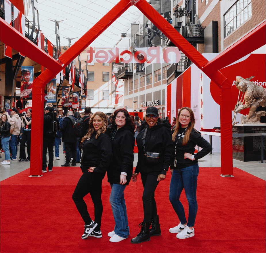 Four smiling women pose together on a red carpet beneath a red Rocket archway at an outdoor event area surrounded by people.