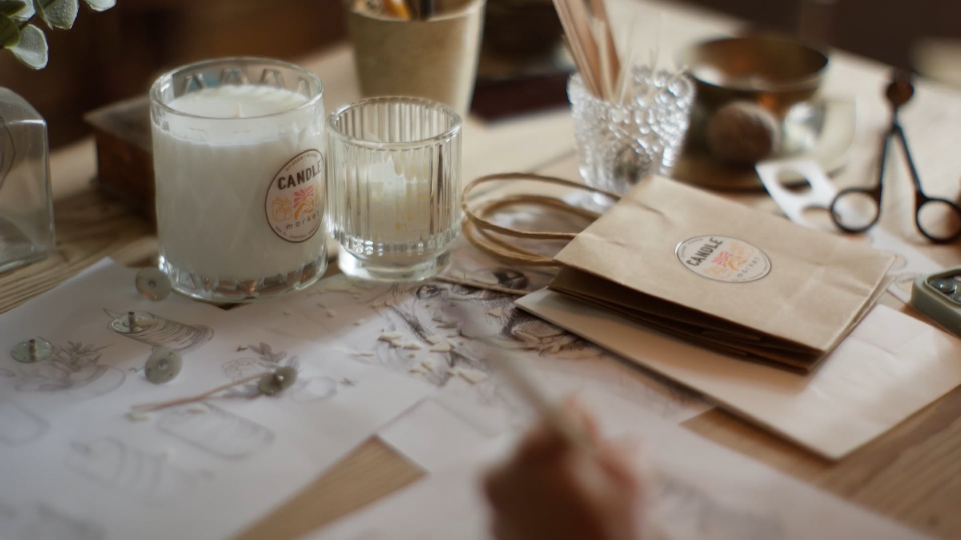 A tabletop workspace with sketches, candles, jars, scissors, and craft supplies, suggesting candle-making.