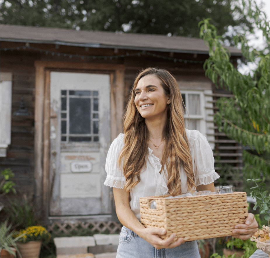 A smiling woman with long hair holding a woven basket outdoors in front of a rustic wooden house.