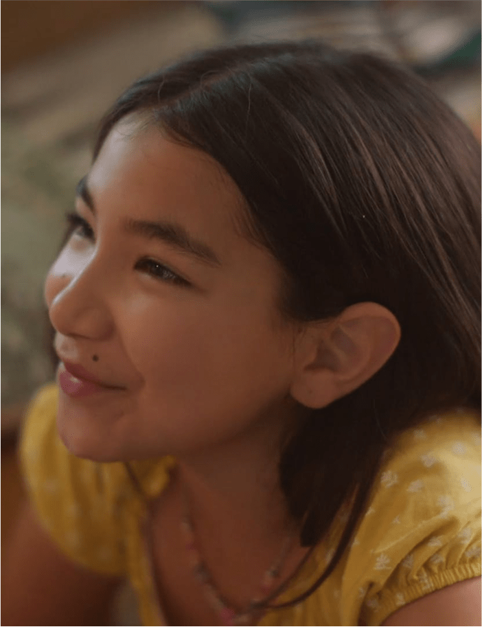 A young girl with dark hair smiling warmly, wearing a yellow top and a colorful beaded necklace.