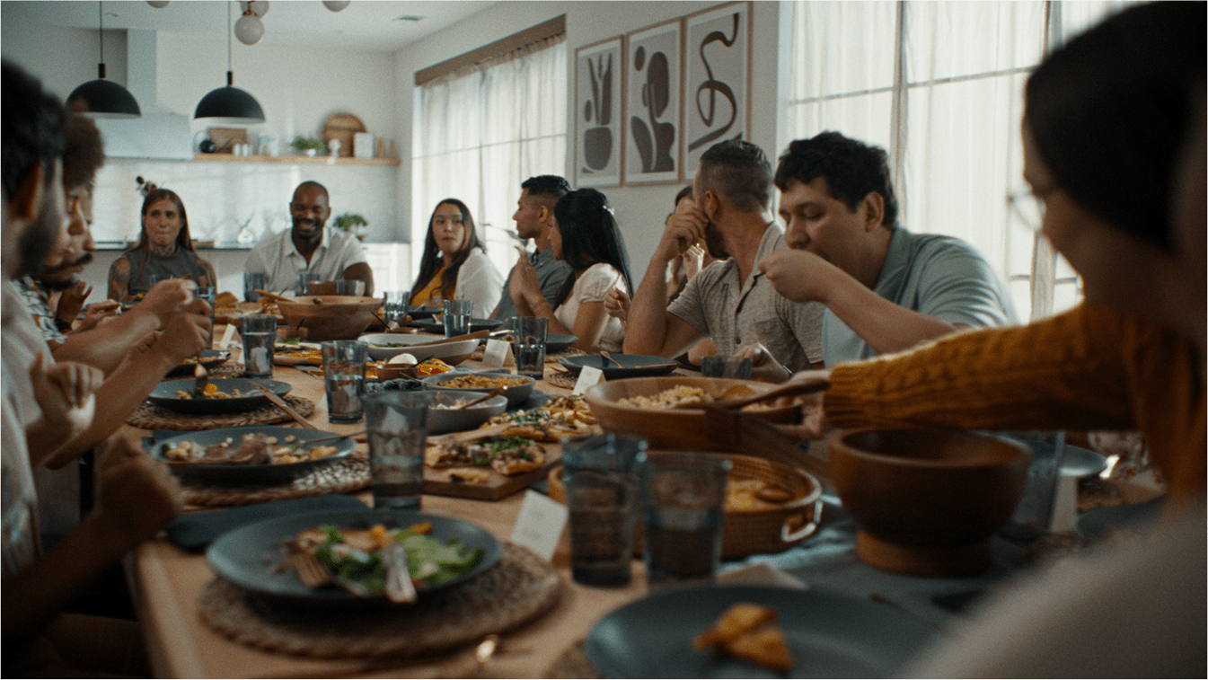 A large group of people sitting around a dining table in a modern, well-lit home, sharing a meal together. The table is set with plates, glasses, and a variety of dishes.