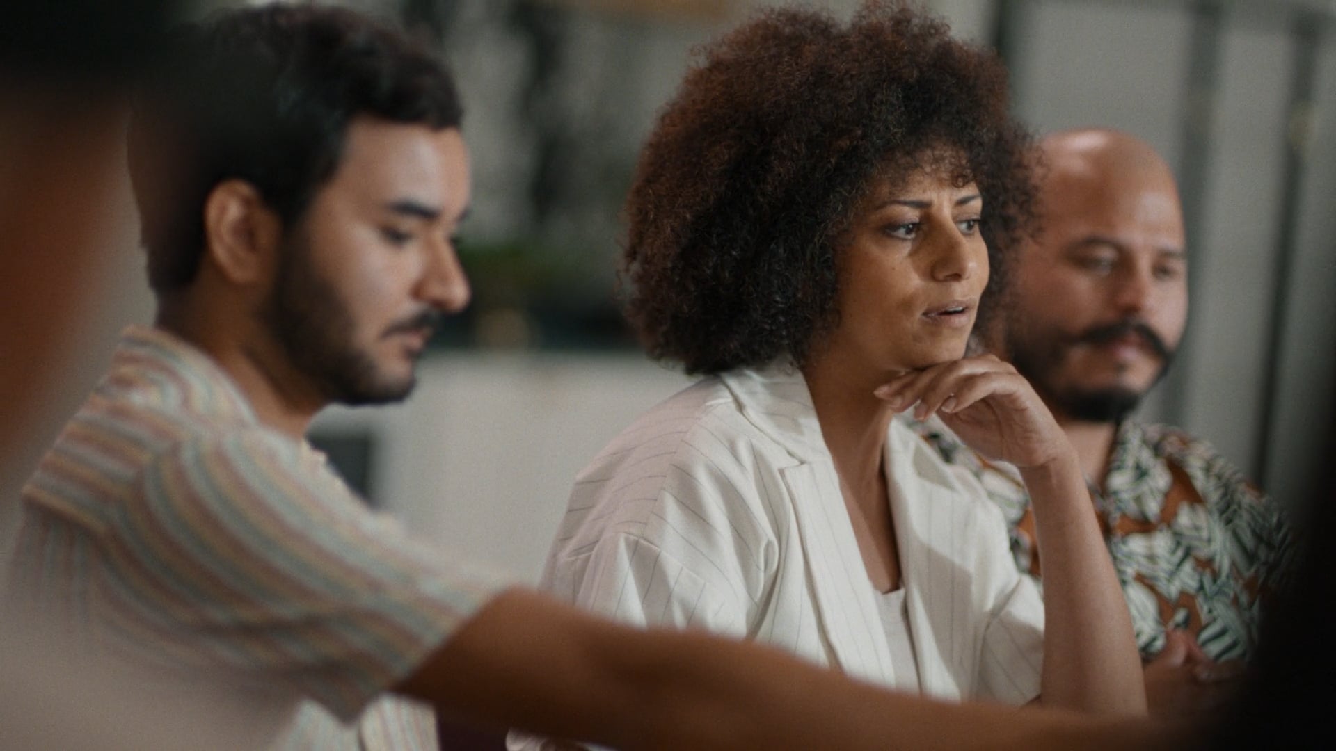 A close-up of a woman with curly hair speaking thoughtfully at the dining table, with two men sitting beside her listening.