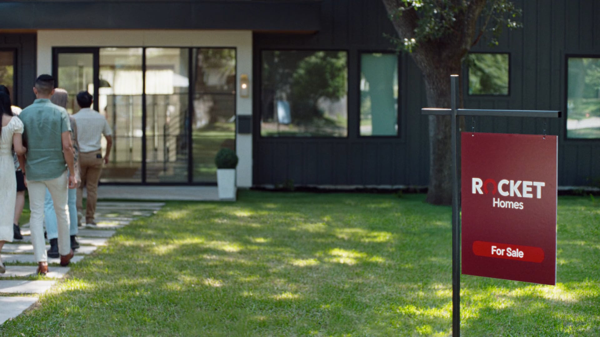 A group of people walking toward a modern black house with large windows. In the foreground, a “Rocket Homes For Sale” sign stands on the lawn.