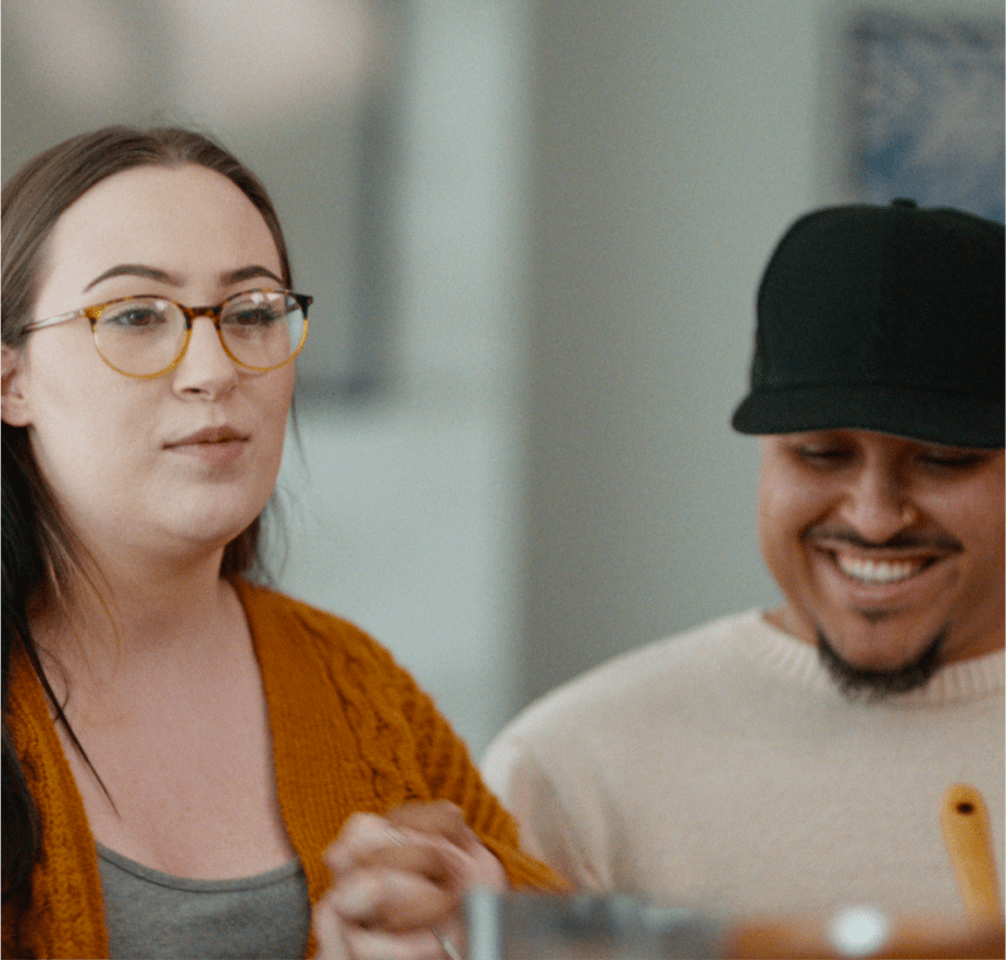 A woman wearing glasses and a mustard-yellow sweater speaks while smiling, sitting next to a man in a cap who is laughing.