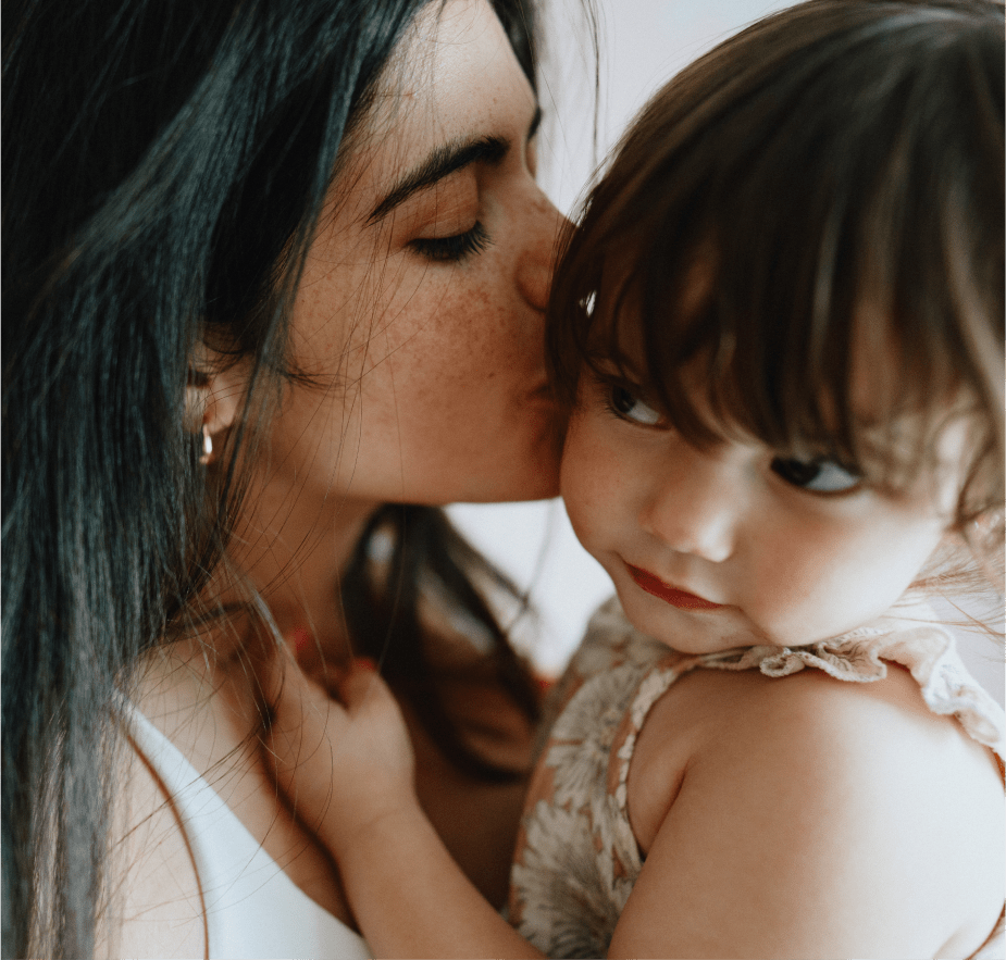 A close-up of a woman tenderly kissing the forehead of a small child she is holding, both with a soft, intimate expression.